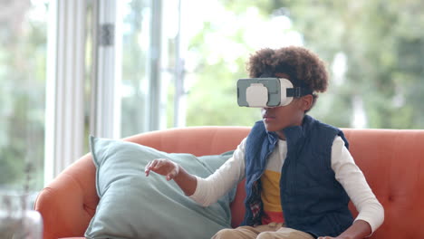 African-american-boy-sitting-on-sofa,-using-vr-headset-and-touching-virtual-screen,-slow-motion