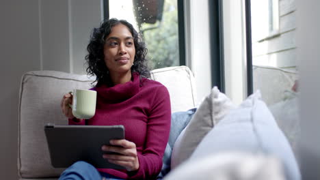 Biracial-woman-sitting-on-sofa-using-tablet-and-drinking-coffee-at-home,-slow-motion