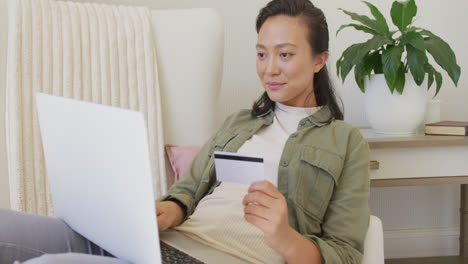 Mujer-Asiática-Feliz-Usando-Una-Computadora-Portátil-Y-Sosteniendo-Una-Tarjeta-De-Crédito-En-El-Dormitorio,-En-Cámara-Lenta