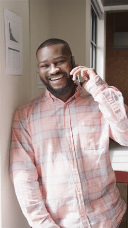 Retrato-En-Vídeo-Vertical-De-Un-Feliz-Hombre-De-Negocios-Afroamericano-Informal-Hablando-Por-Teléfono-Inteligente-En-La-Oficina