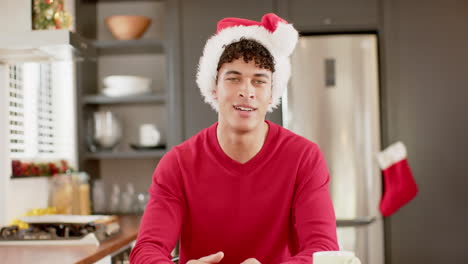 Hombre-Birracial-Con-Sombrero-De-Navidad-En-Videollamada-En-La-Cocina-De-Casa,-Cámara-Lenta,-Espacio-Para-Copiar