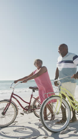 Vertical-video-of-senior-african-american-couple-walking-with-bikes-at-beach,-in-slow-motion