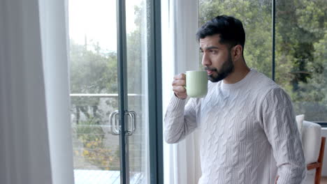 Thoughtful-biracial-man-drinking-coffee-and-looking-through-window-at-home,-slow-motion