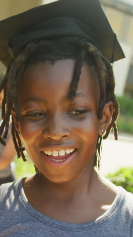 Video-of-happy-african-american-boy-wearing-graduation-hat-and-holding-diploma-in-front-of-school