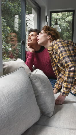 Vertical-video-of-happy-diverse-gay-male-couple-looking-through-window-at-home,-slow-motion