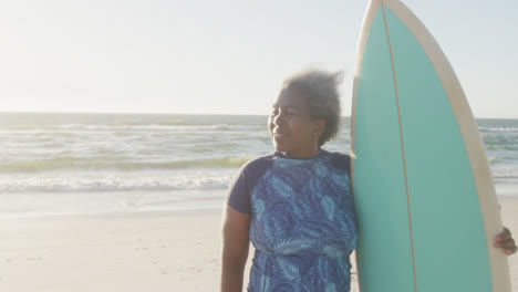 Happy-senior-african-american-woman-holding-surfboard-at-beach,-in-slow-motion,-with-copy-space