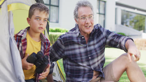 Happy-caucasian-grandfather-and-grandson-sitting-in-tent-and-looking-through-binoculars,-slow-motion