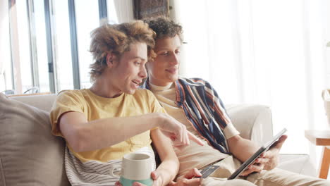Happy-diverse-gay-male-couple-sitting-at-sofa-using-tablet-at-home,-slow-motion