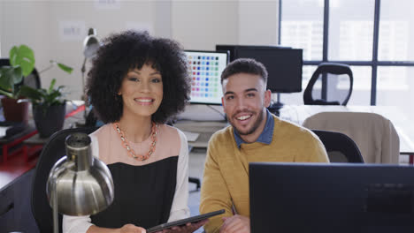 Portrait-of-happy-diverse-male-and-female-colleague-at-desk-using-tablet-and-computer,-slow-motion