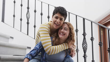 Happy-caucasian-lesbian-couple-sitting-on-stairs,-embracing-and-smiling-in-sunny-house