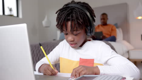 African-american-boy-using-laptop-for-online-lesson-with-father-in-background-at-home,-slow-motion