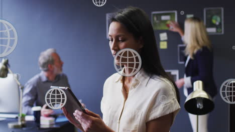 Animation-of-multiple-web-globe-icons-floating-against-biracial-woman-using-digital-tablet-at-office