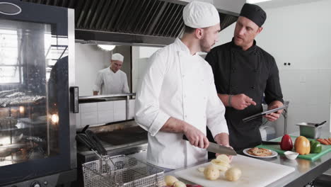 Focused-caucasian-male-chef-instructing-trainee-male-chef-with-tablet-in-kitchen,-slow-motion