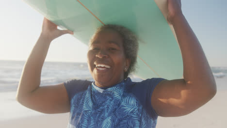 Portrait-of-happy-senior-african-american-woman-holding-surfboard-at-beach,-in-slow-motion