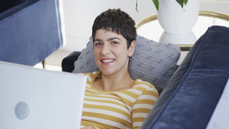 Happy-caucasian-woman-lying-on-sofa,-using-laptop-and-smiling-in-sunny-house