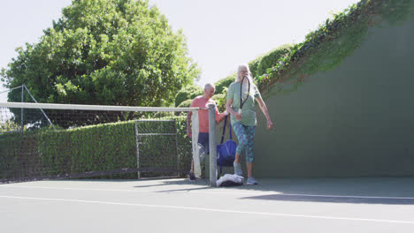 Lächelndes-älteres-Kaukasisches-Paar,-Das-Zum-Tennisspielen-Auf-Einem-Sonnigen-Tennisplatz-Im-Freien-Ankommt,-Zeitlupe