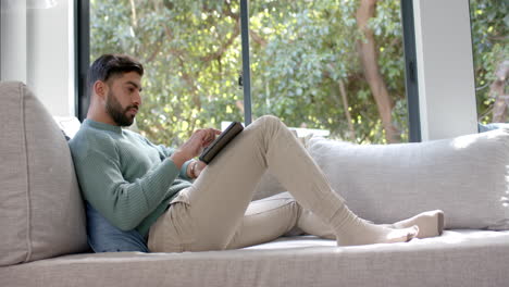 Biracial-man-sitting-on-sofa-using-tablet-at-home,-slow-motion