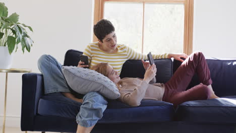 Happy-caucasian-lesbian-couple-lying-on-sofa,-using-tablet-and-smartphone-and-smiling-in-sunny-house