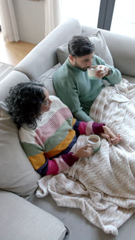 Video-Vertical-De-Una-Pareja-Birracial-Sentada-En-Un-Sofá-Y-Tomando-Café-En-Casa,-Cámara-Lenta