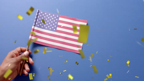 Animation-Von-Konfetti,-Das-über-Eine-Hand-Fällt,-Die-Die-Flagge-Der-Vereinigten-Staaten-Von-Amerika-Auf-Blauem-Hintergrund-Hält