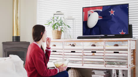 Caucasian-woman-watching-tv-with-rugby-ball-on-flag-of-new-zealand-on-screen