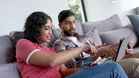 Happy-biracial-couple-sitting-on-sofa-and-using-tablet-for-online-shopping-at-home,-in-slow-motion