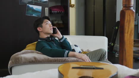 Asian-boy-talking-on-smartphone-sitting-on-the-couch-at-home