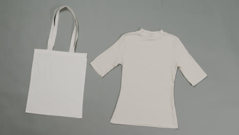 Primer-Plano-De-Bolsa-Blanca-Y-Camiseta-Sobre-Fondo-Gris,-Con-Espacio-Para-Copiar,-Cámara-Lenta