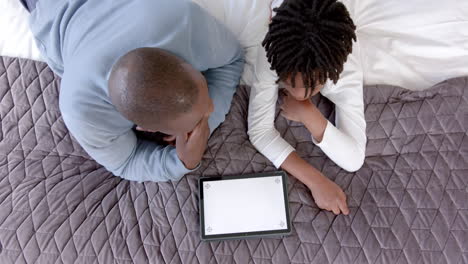 Afroamerikanischer-Vater-Und-Sohn-Liegen-Auf-Dem-Bett-Und-Benutzen-Ein-Tablet-Mit-Platz-Zum-Kopieren-Zu-Hause,-Zeitlupe