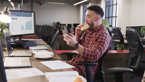 Happy-biracial-casual-businessman-sitting-at-desk,-eating-sandwich-and-using-smartphone,-slow-motion