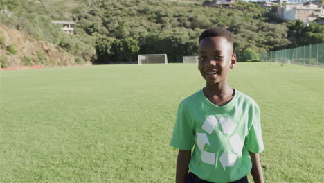 An-African-American-boy-wearing-a-green-recycling-t-shirt-stands-on-a-sunny-soccer-field,-with-copy-