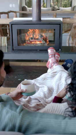 Vertical-video-of-biracial-couple-sitting-on-sofa-in-front-of-fireplace-at-home,-slow-motion