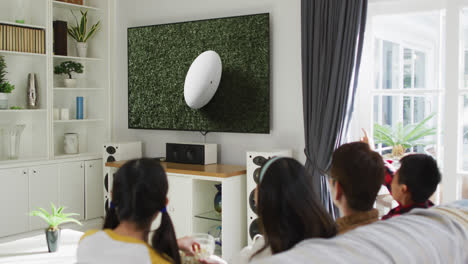 Asian-family-watching-tv-with-rugby-ball-at-stadium-on-screen
