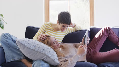 Happy-caucasian-lesbian-couple-lying-on-sofa,-using-tablet,-talking-and-laughing-in-sunny-house