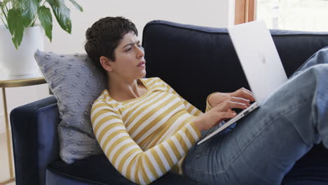 Happy-caucasian-woman-lying-on-sofa,-using-laptop-and-smiling-in-sunny-house