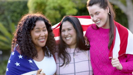 Three-happy-diverse-female-friends-standing-with-flag-on-backs-in-sunny-garden