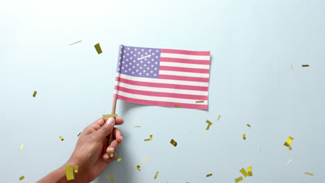 Animation-of-confetti-falling-over-hand-holding-flag-of-united-states-of-america-on-blue-background