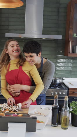 Feliz-Lesbiana-Caucásica-Preparando-Comida,-Abrazando-Y-Usando-Tableta-En-La-Soleada-Cocina