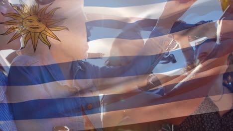 Composite-Video-Einer-Wehenden-Uruguayischen-Flagge-über-Einer-Gruppe-Unterschiedlicher-Freunde,-Die-Im-Auto-Mit-Bier-Anstoßen