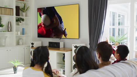 Asian-family-watching-tv-with-diverse-male-rugby-players-playing-on-screen
