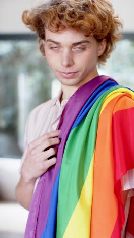 Vertikales-Video-Eines-Porträts-Eines-Glücklichen-Kaukasischen-Mannes,-Der-Zu-Hause-Die-Regenbogen-LGBT-Flagge-Hält,-Zeitlupe