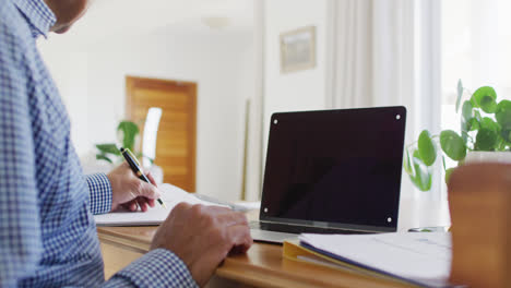 Video-Eines-älteren-Mannes-Mit-Gemischter-Abstammung,-Der-Zu-Hause-Am-Schreibtisch-Sitzt,-Schreibt-Und-Einen-Laptop-Verwendet,-Platz-Auf-Dem-Bildschirm-Kopieren