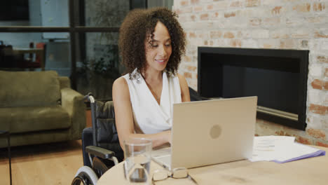 Happy-biracial-businesswoman-in-wheelchair-using-laptop-at-desk-in-office,-in-slow-motion