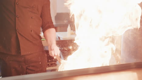 Caucasian-male-chef-frying-food-in-frying-pan-with-bursting-fire-in-kitchen,-slow-motion