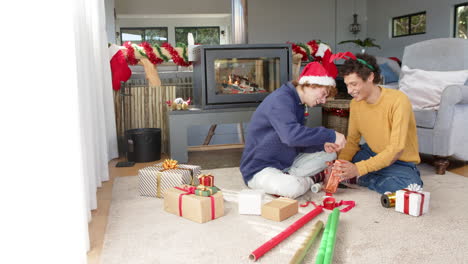 Diverse-gay-male-couple-wrapping-christmas-presents-by-fireplace-at-home,-slow-motion,-copy-space
