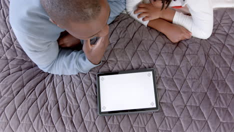 Afroamerikanischer-Vater-Und-Sohn-Liegen-Auf-Dem-Bett-Und-Benutzen-Ein-Tablet-Mit-Platz-Zum-Kopieren-Zu-Hause,-Zeitlupe