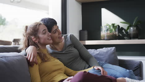 Happy-caucasian-lesbian-couple-sitting-on-sofa,-embracing-and-talking-in-sunny-living-room