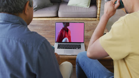 Biracial-father-and-son-watching-laptop-with-african-american-male-rugby-player-with-ball-on-screen