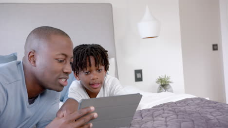 Glücklicher-Afroamerikanischer-Vater-Und-Sohn-Liegen-Zu-Hause-Mit-Tablet-Im-Bett,-Zeitlupe,-Kopierraum