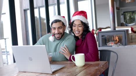 Glückliches-Gemischtrassiges-Paar-Mit-Weihnachtsmannmützen,-Das-Zu-Hause-In-Zeitlupe-Einen-Laptop-Für-Videoanrufe-Verwendet